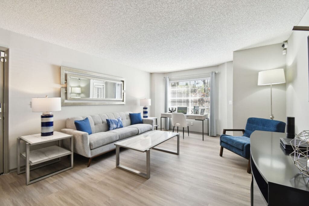 Dog-Friendly Apartments In Lake Underhill, FL - Enclave At Lake Underhill - Living Room Area With Wood-Style Flooring, Couch, Small Tables with Lamps,Desk, And A Large Window with Blinds and Curtains.